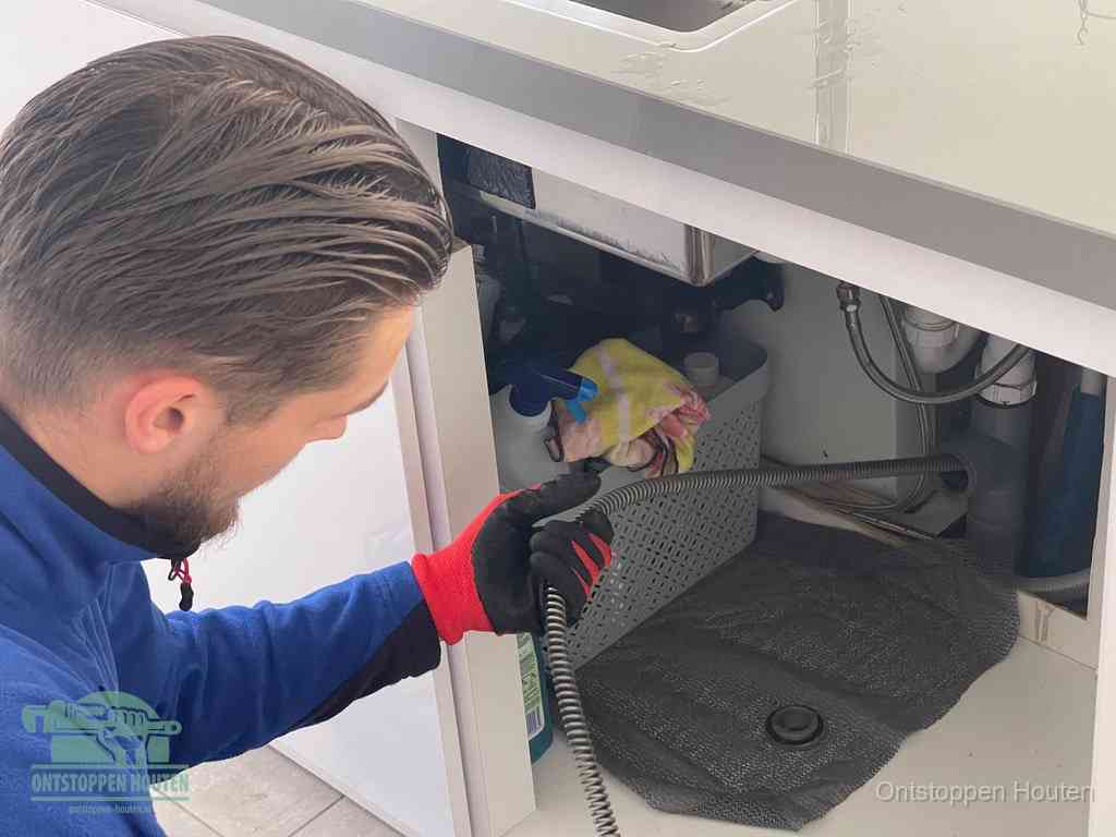 Een monteur voert een flexibele ontstoppingsveer in onder een keukenkastje om een verstopping voorbij de sifon te bereiken in Houten