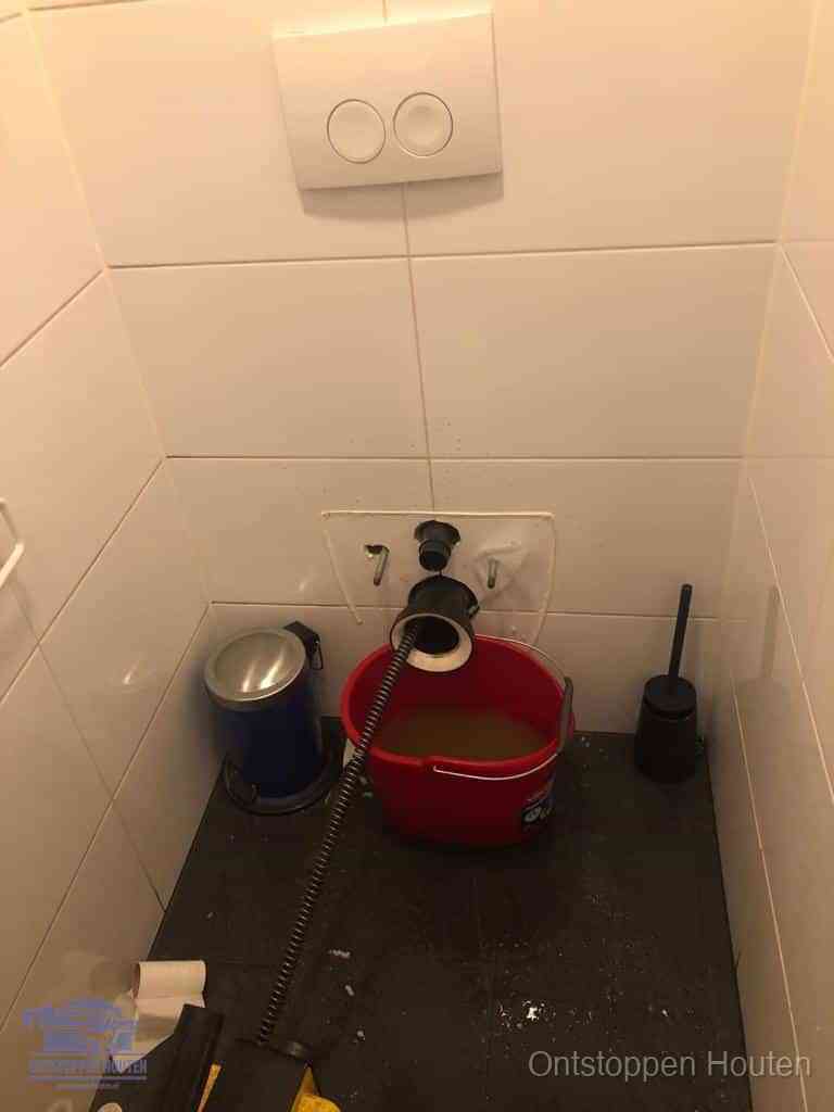 Een ontstopte wand-wc met verwijderde toiletpot waar een ontstoppingsveer in de wanduitgang verdwijnt, het achterhalen waar de verstopping zit in Houten