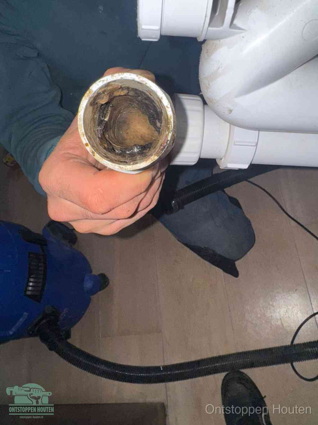 Een hand houdt een losgekoppeld PVC-afvoerbuisstuk vast dat van binnen volledig is dichtgeslibd met vet, vuil en aanslag, de meest voorkomende oorzaak van een verstopte afvoer in Houten
