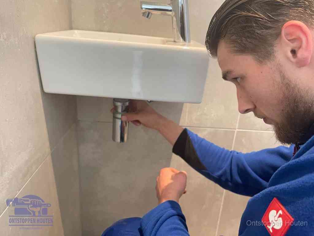 Een monteur draait met de hand de zwanenhalssifon onder een fonteinwasbak in de toiletruimte los om de bron van rioollucht in Houten te vinden
